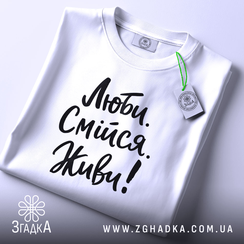 Футболка люби смійся живи, білого кольору, складена на плоскій поверхні, фон світло-сірий, принт чорного кольору з написом 'Люби. Смійся. Живи!' у стилі рукописного шрифту, з етикеткою зеленого кольору.