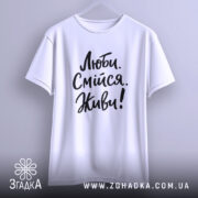 Футболка люби смійся живи, білого кольору, підвішена на вішалці, фон світло-сірий, принт чорного кольору з написом 'Люби. Смійся. Живи!' у стилі рукописного шрифту.