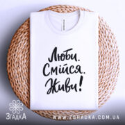 Футболка люби смійся живи, білого кольору, складена на круглій кошику, фон світло-сірий, принт чорного кольору з написом 'Люби. Смійся. Живи!' у стилі рукописного шрифту.