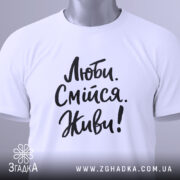 Футболка люби смійся живи, білого кольору, складена на плоскій поверхні, фон світло-сірий, принт чорного кольору з написом 'Люби. Смійся. Живи!' у стилі рукописного шрифту, з етикеткою зеленого кольору.