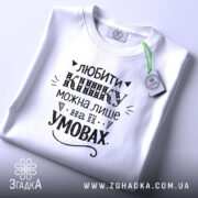 Футболка любити кішку білого кольору, складена на поверхні, зображена з верхнього ракурсу, фон світло-сірий, принт чорного кольору з написом та етикеткою з зеленим шнурком.