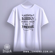 Футболка любити кішку білого кольору, підвішена на вішалці, зображена з фронтального ракурсу, фон світло-сірий, принт чорного кольору з написом та декоративними елементами.