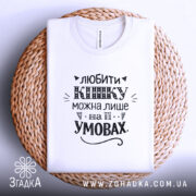 Футболка любити кішку білого кольору, складена на плетеній основі, зображена з верхнього ракурсу, фон білий, принт чорного кольору з написом та декоративними елементами.