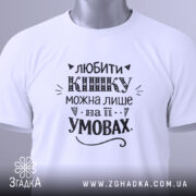 Футболка любити кішку білого кольору, складена на поверхні, зображена з верхнього ракурсу, фон світло-сірий, принт чорного кольору з написом та етикеткою з зеленим шнурком.
