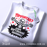 Футболка лови віртуальні обійми, біла, зображена зверху, складена на плоскій поверхні, фон світло-сірий, принт з черепом, що розпростерті руки, червоні сердечка та текст "Прочитав? Лови віртуальні обійми!", на етикетці зелена стрічка.