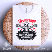 Футболка лови віртуальні обійми, біла, зображена зверху, складена на плоскій поверхні, фон світло-сірий, принт з черепом, що розпростерті руки, червоні сердечка та текст "Прочитав? Лови віртуальні обійми!".