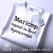 Футболка магістр чорної бухгалтерії, білого кольору, складена на поверхні, фон світло-сірий, принт чорного кольору з текстом 'Магістр чорної та білої бухгалтерії', на комірці видна етикетка.