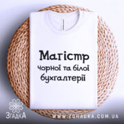 Футболка магістр чорної бухгалтерії, білого кольору, складена на плетеній тарілці, фон світло-сірий, принт чорного кольору з текстом 'Магістр чорної та білої бухгалтерії'.