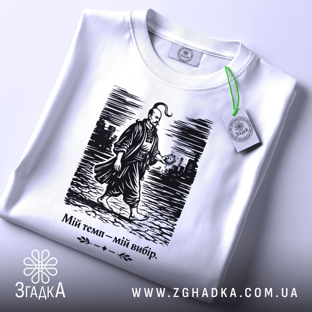 Футболка мій темп мій вибір. Біла футболка, зображена зверху, з коротким рукавом, на ній принт з чоловіком у традиційному одязі, що йде по вулиці. Принт чорного кольору, текст під ним також чорний. На футболці видно етикетку з зеленим шнурком.