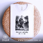 Футболка мій темп мій вибір. Біла футболка, складена на плетеній кошику. Принт з чоловіком у традиційному одязі, що йде по вулиці, виконаний в чорному кольорі. Текст під зображенням також чорний. Фон світлий, з плетеним текстурованим елементом.