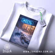 Футболка міська жесть, біла, складена на столі, фон світло-сірий, принт з зображенням людини, що лежить у снігу з лопатою, на фоні магазину з неоновим написом 'ЖЕСТЬ', на футболці є етикетка з зеленим шнурком.