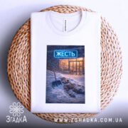 Футболка міська жесть, біла, складена на кошику, фон світло-сірий, принт з зображенням людини, що лежить у снігу з лопатою, на фоні магазину з неоновим написом 'ЖЕСТЬ'.
