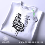 Футболка мої дівчата сила. Складена біла футболка з принтом чорного кольору, на якому напис "Мої дівчата = моя сила" та зображення жінок. На футболці є етикетка з зеленим шнурком. Фон світло-сірий.