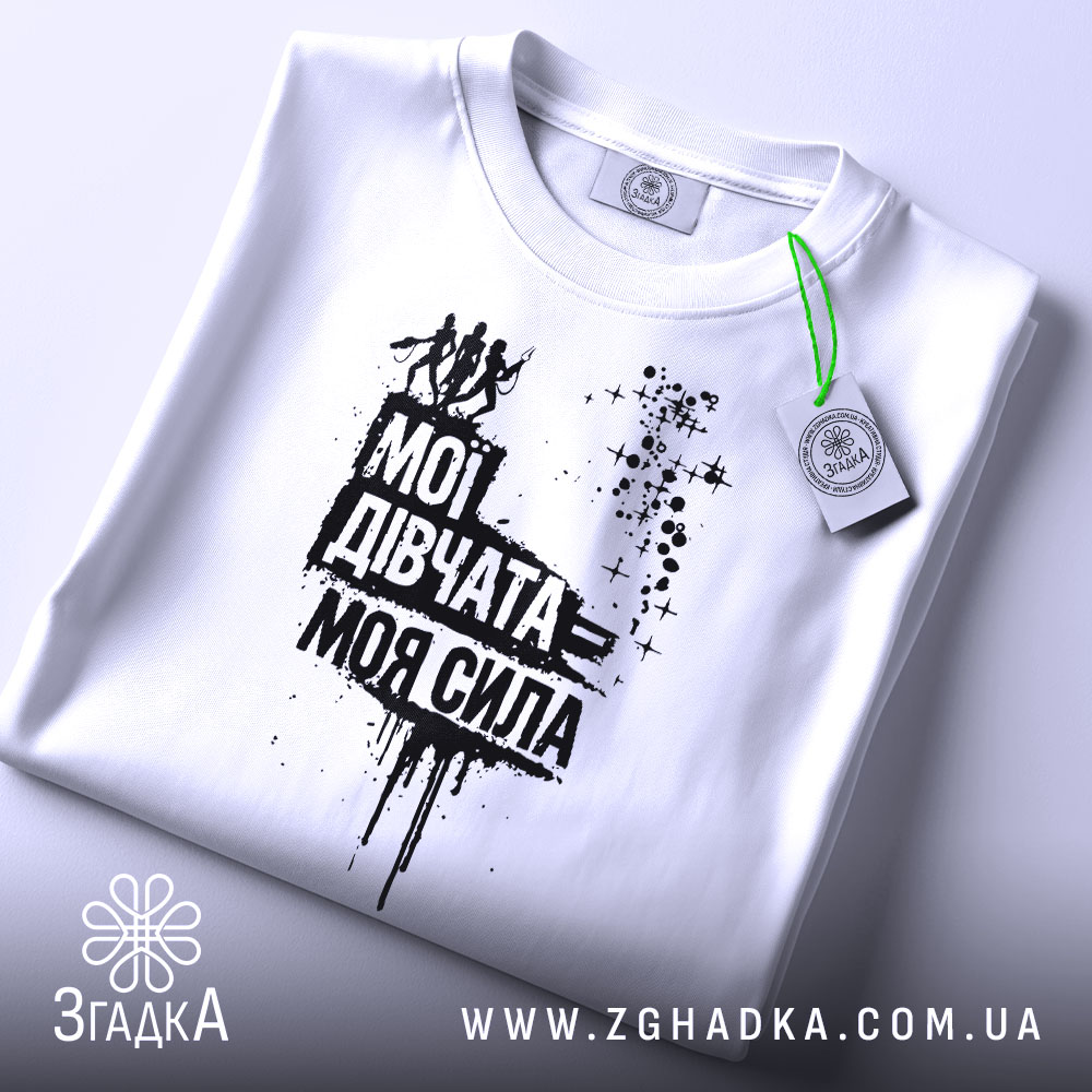 Футболка мої дівчата сила. Складена біла футболка з принтом чорного кольору, на якому напис "Мої дівчата = моя сила" та зображення жінок. На футболці є етикетка з зеленим шнурком. Фон світло-сірий.