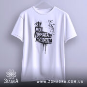 Футболка мої дівчата сила. Біла футболка, підвішена на вішалці, з принтом чорного кольору, що містить текст "Мої дівчата = моя сила" та зображення жінок. Фон світло-сірий, деталі принта виконані у стилі графіті.