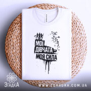 Футболка мої дівчата сила. Складена біла футболка на плетеній основі, з принтом чорного кольору, що містить текст "Мої дівчата = моя сила" та зображення жінок. Фон білий, деталі принта виконані у стилі графіті.