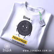 Футболка моя краща половинка, білого кольору, складена на столі, фон світло-сірий, принт з персонажем у вигляді печива Oreo, напис 'Вона - моя краща половинка' жовтого кольору, на футболці є етикетка з зеленим шнурком.