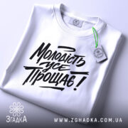 Футболка молодість усе прощає. Білий колір, зображена зверху на плоскій поверхні, фон світло-сірий. Принт чорного кольору з написом "Молодість усе прощає!" у стилі рукописного шрифту, з зеленим ярликом.