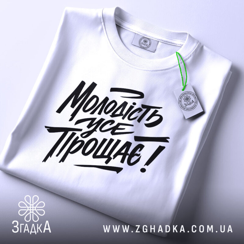 Футболка молодість усе прощає. Білий колір, зображена зверху на плоскій поверхні, фон світло-сірий. Принт чорного кольору з написом "Молодість усе прощає!" у стилі рукописного шрифту, з зеленим ярликом.