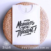 Футболка молодість усе прощає. Білий колір, зображена зверху на плоскій поверхні, фон світло-сірий. Принт чорного кольору з написом "Молодість усе прощає!" у стилі рукописного шрифту, на плетеній основі.