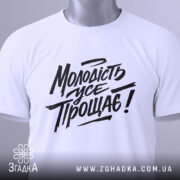 Футболка молодість усе прощає. Білий колір, зображена зверху на плоскій поверхні, фон світло-сірий. Принт чорного кольору з написом "Молодість усе прощає!" у стилі рукописного шрифту, з зеленим ярликом.