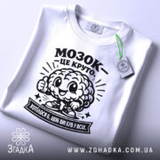 Футболка мозок це круто. Біла футболка, зображена зверху, складена на поверхні. На футболці чорний принт з мозком, що усміхається, та написом 'МОЗОК - ЦЕ КРУТО'. На комірці видно етикетку з зеленим ярликом. Фон світлий, з легким градієнтом.