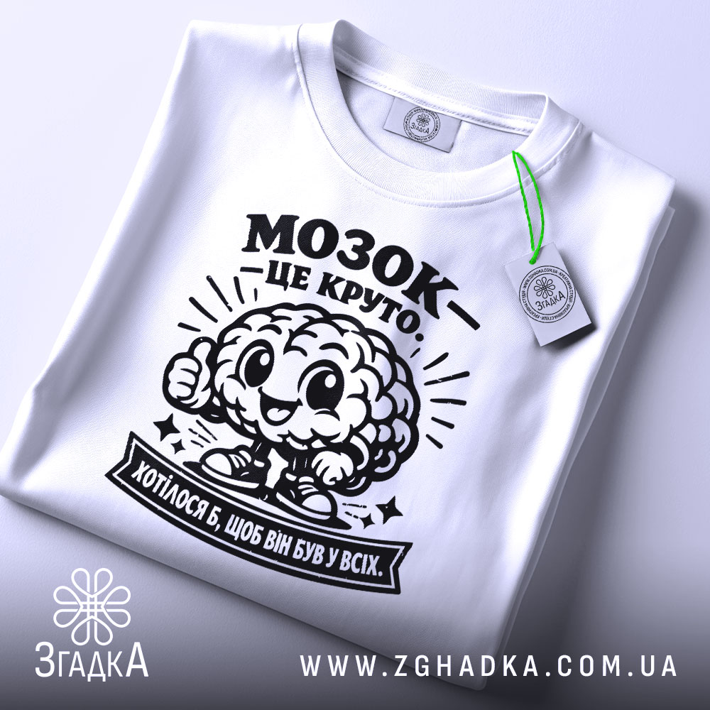Футболка мозок це круто. Біла футболка, зображена зверху, складена на поверхні. На футболці чорний принт з мозком, що усміхається, та написом 'МОЗОК - ЦЕ КРУТО'. На комірці видно етикетку з зеленим ярликом. Фон світлий, з легким градієнтом.