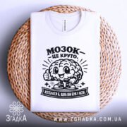 Футболка мозок це круто. Біла футболка, зображена зверху, складена на плетеній кошику. На футболці чорний принт з усміхненим мозком та написом 'МОЗОК - ЦЕ КРУТО'. Фон світлий, з плетеним текстурованим елементом.