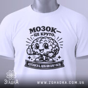 Футболка мозок це круто. Біла футболка, зображена зверху, складена на поверхні. На футболці чорний принт з мозком, що усміхається, та написом 'МОЗОК - ЦЕ КРУТО'. На комірці видно етикетку з зеленим ярликом. Фон світлий, з легким градієнтом.