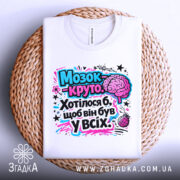 Футболка мозок це круто. Біла футболка, розкладена на плетеній основі, з яскравим принтом, що містить мозок і текст. Принт виконаний у рожевих і блакитних кольорах, фон нейтральний.