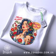 Футболка мила як рай. Біла футболка, складена на столі, з принтом жінки з синім волоссям. На футболці є етикетка з зеленим шнурком. Фон світлий, акцент на принті.