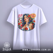 Футболка мила як рай. Біла футболка, підвішена на вішалці, з яскравим принтом жінки з синім волоссям. Фон світлий, акцент на принті.