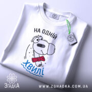 Футболка на одній хвилі, білого кольору, складена на столі, фон світло-сірий, принт з персонажем у червоному краватці та написом 'На одній хвилі' у блакитному кольорі, з зеленим ярликом.