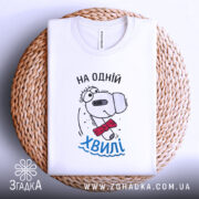 Футболка на одній хвилі, білого кольору, складена на плетеному кошику, фон білий, принт з персонажем у червоному краватці та написом 'На одній хвилі' у блакитному кольорі.