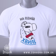 Футболка на одній хвилі, білого кольору, складена на столі, фон світло-сірий, принт з персонажем у червоному краватці та написом 'На одній хвилі' у блакитному кольорі, з зеленим ярликом.