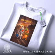 Футболка на роботу білого кольору, розкладена на поверхні, фон світлий, принт з двома персонажами, один у костюмі клоуна, інший у зеленому капелюсі, текст на принті: 'НА РОБОТУ – ЯК НА СВЯТО!', на футболці є етикетка з зеленим шнурком.