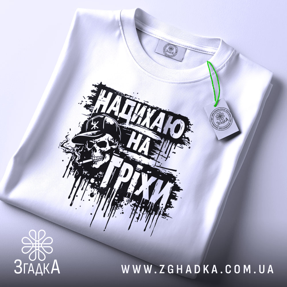 Футболка надихаю на гріхи, біла, складена на поверхні, фон світлий, принт чорного кольору з черепом у капелюсі, прикріплена етикетка з зеленим шнурком, виглядає елегантно.