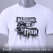 Футболка надихаю на гріхи, біла, складена на поверхні, фон світлий, принт чорного кольору з черепом у капелюсі, прикріплена етикетка з зеленим шнурком, виглядає елегантно.