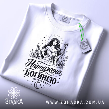 Футболка Народжена богинею білого кольору, складена на плоскій поверхні, фон світло-сірий, принт з богинею в довгій сукні, що тримає жезл, з елементами рослинності та зірками, з етикеткою на зеленій стрічці.