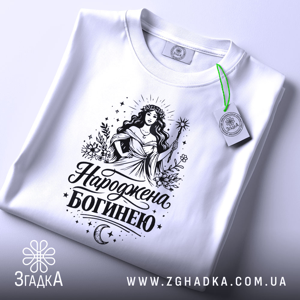 Футболка Народжена богинею білого кольору, складена на плоскій поверхні, фон світло-сірий, принт з богинею в довгій сукні, що тримає жезл, з елементами рослинності та зірками, з етикеткою на зеленій стрічці.