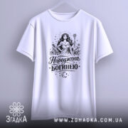 Футболка Народжена богинею білого кольору, підвішена на вішалці, фон світло-сірий, принт з богинею в довгій сукні, що тримає жезл, оточена рослинними елементами та зірками.