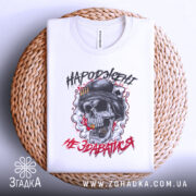 Футболка народжені не здаватися, білого кольору, зображена зверху на плетеній кошику, фон світло-сірий, принт з черепом у військовій касці, оточений димом та червоними акцентами.