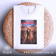 Футболка навколо хаос. Біла футболка, складена на плетеній основі, з принтом, що зображує пару на фоні міста. Принт яскравий, з червоним і блакитним текстом, що читається: 'Навколо хаос. Між нами — спокій.'