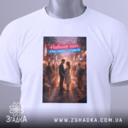 Футболка навколо хаос. Біла футболка, складена на поверхні, з принтом, що зображує пару в обіймах на фоні міста. Принт яскравий, з червоним і блакитним текстом, що читається: 'Навколо хаос. Між нами — спокій.' На футболці є етикетка з зеленим шнурком.