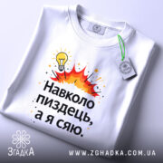 Футболка навколо пиздець, біла, складена на поверхні, фон світло-сірий, принт з жовтою лампочкою та червоними вибухами, текст чорного кольору, з етикеткою на комірці.