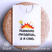 Футболка навколо пиздець, біла, складена на плетеній кошику, фон світло-сірий, принт з жовтою лампочкою та червоними вибухами, текст чорного кольору.