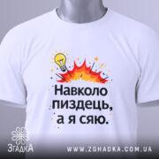 Футболка навколо пиздець, біла, складена на поверхні, фон світло-сірий, принт з жовтою лампочкою та червоними вибухами, текст чорного кольору, з етикеткою на комірці.