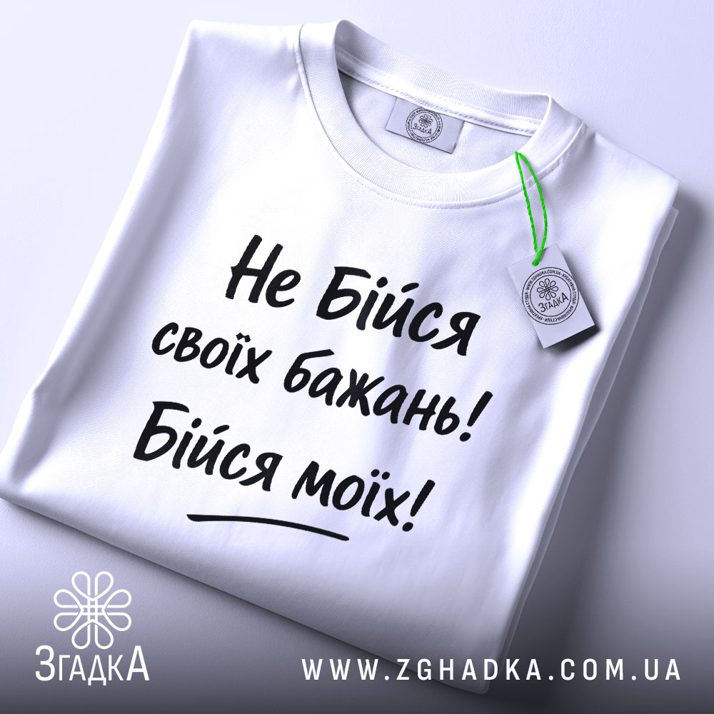 Футболка не бійся бажань. Біла футболка, складена на поверхні, з видимим етикеткою, фон світло-сірий, принт чорного кольору з текстом "Не Бійся своїх бажань! Бійся моїх!".
