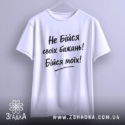 Футболка не бійся бажань. Біла футболка з коротким рукавом, підвішена на вішалці, фон світло-сірий, принт чорного кольору з текстом "Не Бійся своїх бажань! Бійся моїх!".