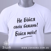 Футболка не бійся бажань. Біла футболка, складена на поверхні, з видимим етикеткою, фон світло-сірий, принт чорного кольору з текстом "Не Бійся своїх бажань! Бійся моїх!".